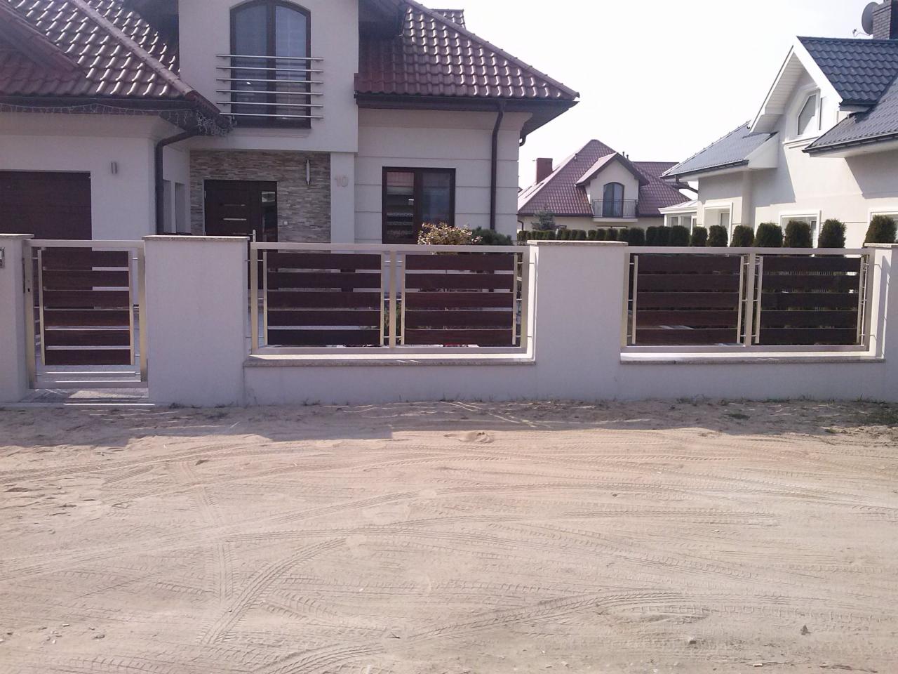 Nowoczesne ogrodzenie posesji z drewnianymi panelami i metalowymi słupkami na tle jasnego domu jednorodzinnego z brązowym dachem i zadbanym trawnikiem.