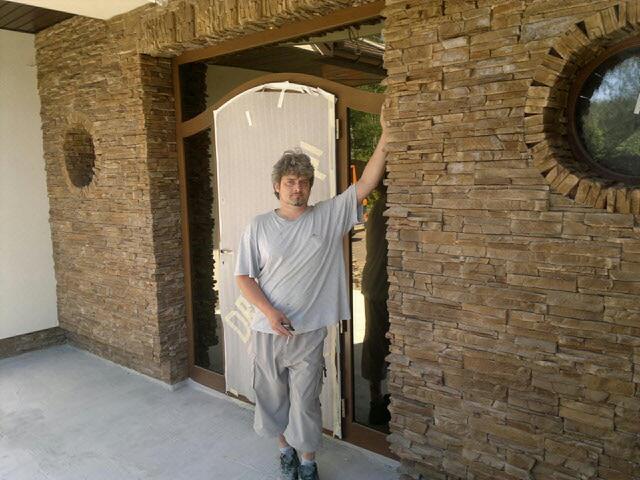 Elewacja budynku wykończona kamieniem dekoracyjnym, z widocznym oknem w kształcie koła i mężczyzną opierającym się o ścianę. Naturalne odcienie brązu i beżu kamienia.