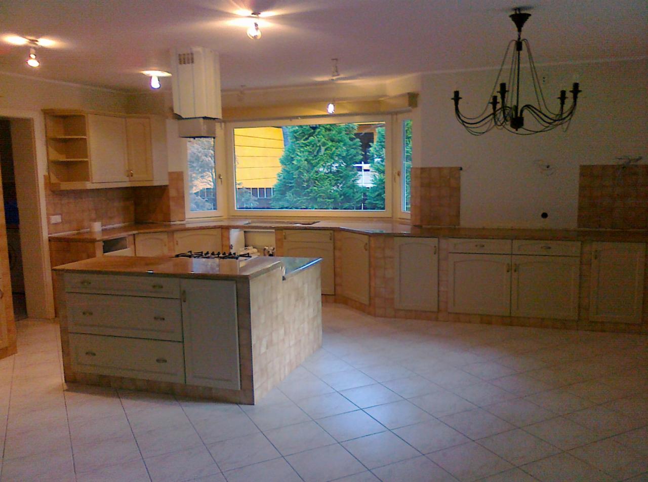 Jasna kuchnia w stylu retro z wyspą, kremowymi szafkami i brązowymi płytkami. Duże okno z widokiem na zieleń. Elementy dekoracyjne, jak żyrandol, dodają charakteru.