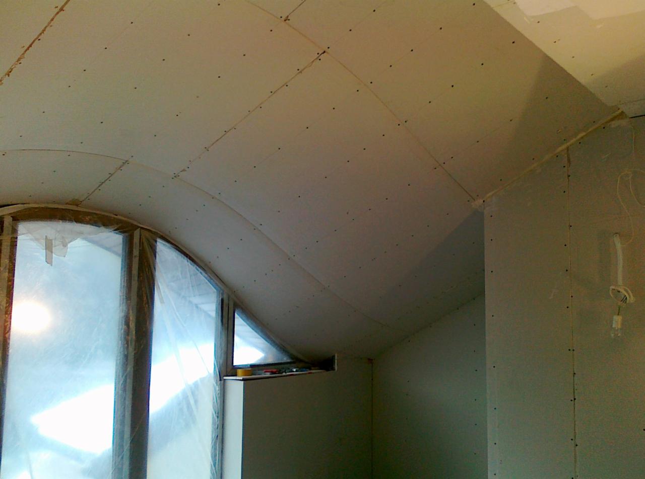 Montaż płyt karton-gips na zakrzywionym suficie poddasza z oknem w kształcie łuku. Widoczne śruby mocujące płyty i fragment ściany bocznej. Prace wykończeniowe.