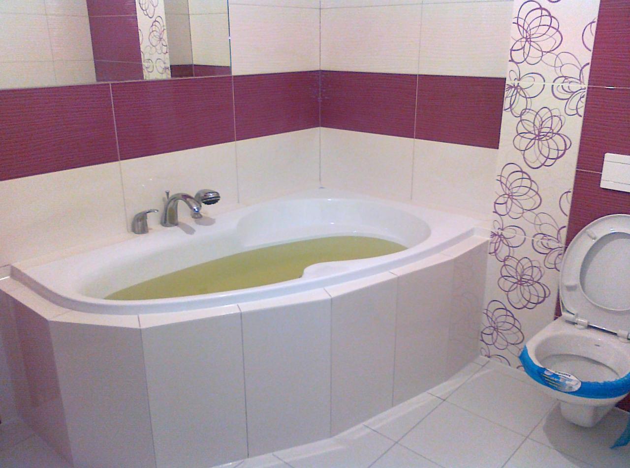 Wykończona łazienka z narożną wanną wypełnioną wodą, białe i fioletowe płytki, oraz toaleta z niebieskim akcentem. W tle widoczne lustro.