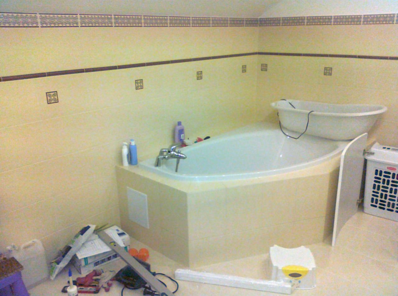 Wykończona, jasna łazienka z narożną wanną obudowaną płytkami, dekoracyjnym paskiem płytek na ścianie i akcesoriami. Widoczne narzędzia glazurnicze.