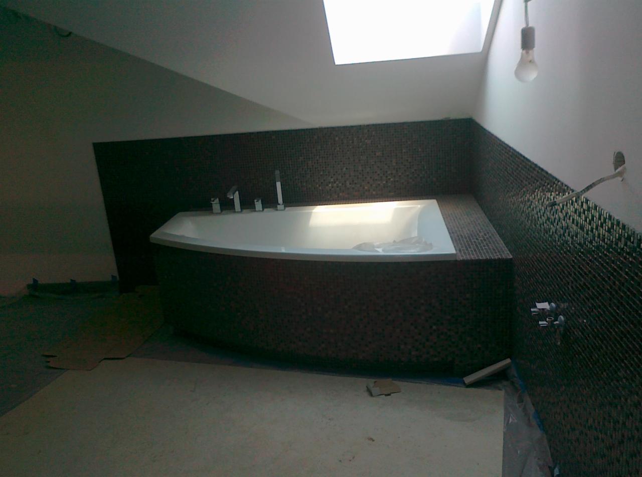 Wanna obudowana ciemną mozaiką w łazience na poddaszu z oknem dachowym. Widoczne niedokończone prace wykończeniowe i wystająca żarówka.