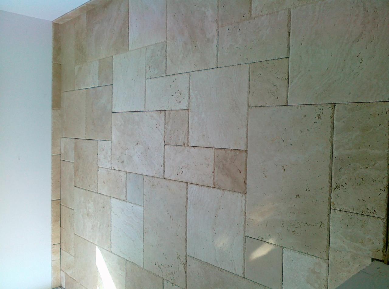 Ściana wyłożona beżowymi, porowatymi płytkami w różnych rozmiarach, ułożonymi w nieregularny wzór. Widoczny fragment białej ściany z lewej strony kadru.