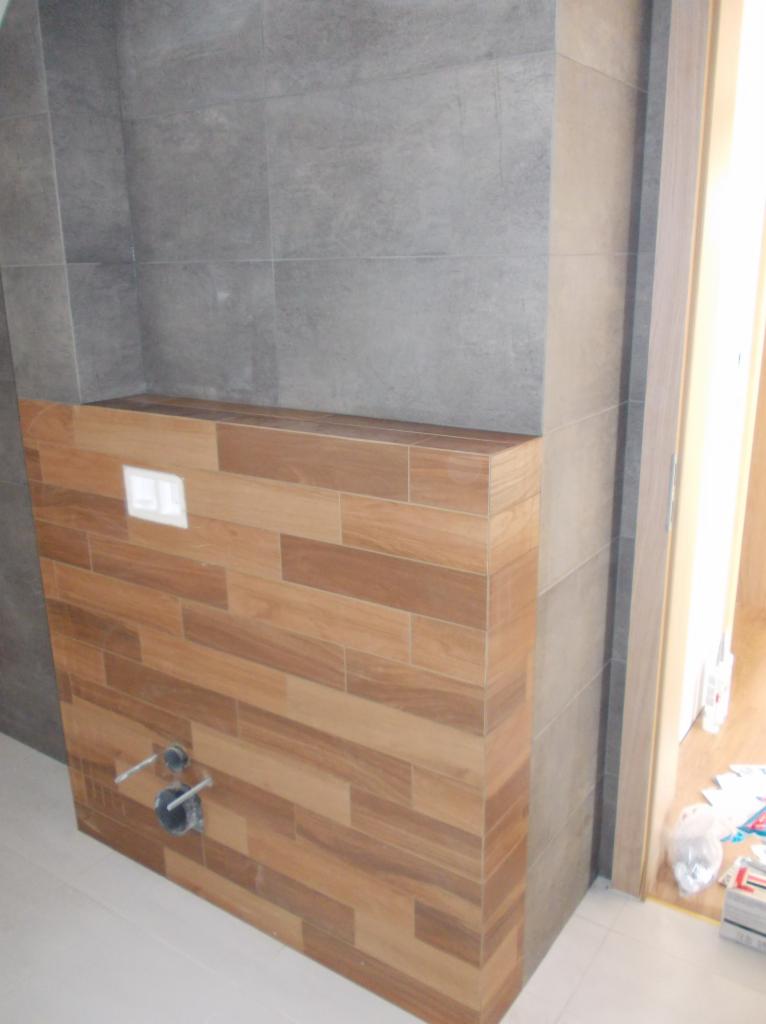 Łazienka w trakcie wykończenia: drewnopodobne płytki na zabudowie stelaża WC, szare płytki na ścianie, widoczne przyłącza wodne. Montaż podtynkowy.