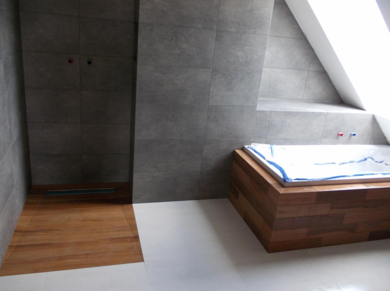 Nowoczesna łazienka na poddaszu: szare płytki ścienne, drewnopodobna strefa prysznicowa z odpływem liniowym i obudowana drewnem wanna z zabezpieczeniem.