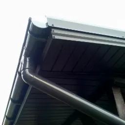 Szary system rynnowy zamontowany na dachu z drewnianą podbitką, widok z dołu pod kątem. Widoczne mocowania rynny i fragment drzewa w lewym górnym rogu.