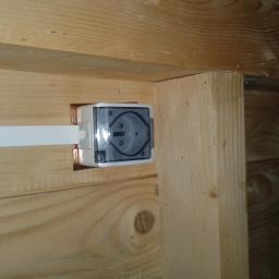 Instalacje-techniczne.pl - Zamontowane gniazdko elektryczne z przezroczystą osłoną w drewnianej ścianie. Widoczny kabel zasilający. Ujęcie z bliska na tle drewnianej konstrukcji.