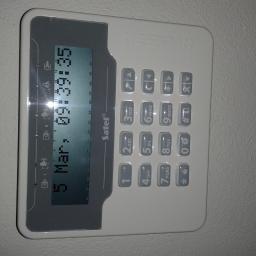 Instalacje-techniczne.pl - Panel sterowania alarmem Sater z wyświetlaczem LCD pokazującym datę i godzinę, umieszczony na jasnej ścianie. Widoczne przyciski numeryczne i funkcyjne.