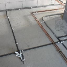 Instalacje-techniczne.pl - Instalacja podłogowa: rury miedziane i czarne peszle przymocowane do surowego betonu, przygotowane do zalania posadzką. Widoczny fragment ściany z betonu komórkowego.