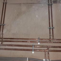 Instalacje-techniczne.pl - Miedziane rury instalacji centralnego ogrzewania, zamocowane do surowej ściany. Widoczne złączki i uchwyty mocujące. Szare rury karbowane w dolnej części kadru.
