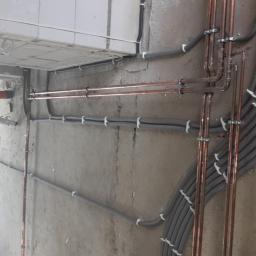 Instalacje-techniczne.pl - Miedziane i szare rury instalacyjne prowadzone wzdłuż surowej ściany, z widocznymi mocowaniami i fragmentem okna. Instalacje w nowym budynku.