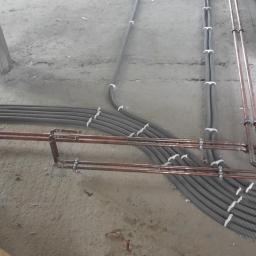 Instalacje-techniczne.pl - Szczegółowe ułożenie rur miedzianych i peszli instalacji elektrycznej na surowym podłożu wewnątrz budynku, widoczne elementy mocujące.