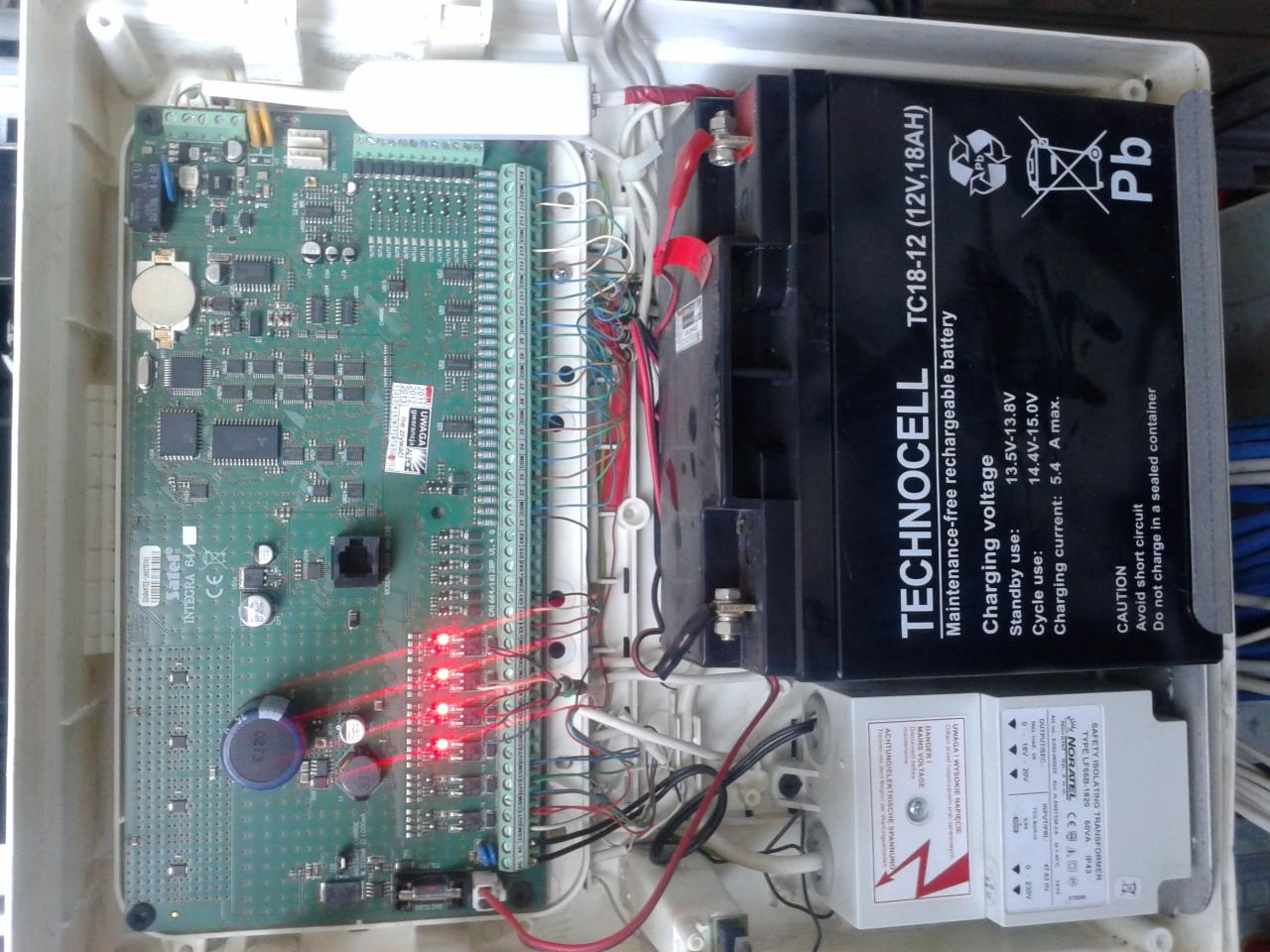 Otwarta obudowa centrali alarmowej z widoczną elektroniką, akumulatorem i transformatorem. Czerwone diody LED sygnalizują działanie systemu.