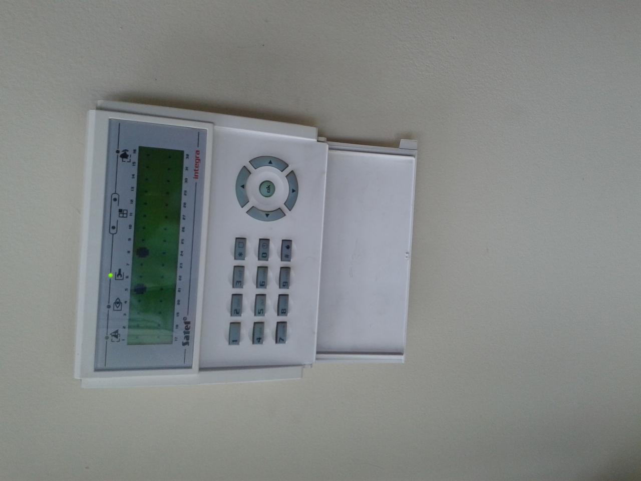 Biały panel sterowania alarmem firmy Satel Integra z otwartą klapką, zamontowany na jasnej ścianie. Widoczny wyświetlacz LCD i klawiatura numeryczna.
