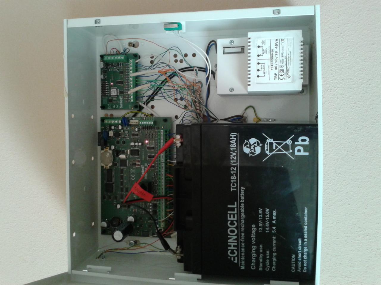 Otwarta skrzynka sterownicza z elektroniką, akumulatorem i transformatorem. Widoczne przewody, płytki drukowane i elementy zasilania.  Szczegółowe ujęcie wnętrza instalacji.