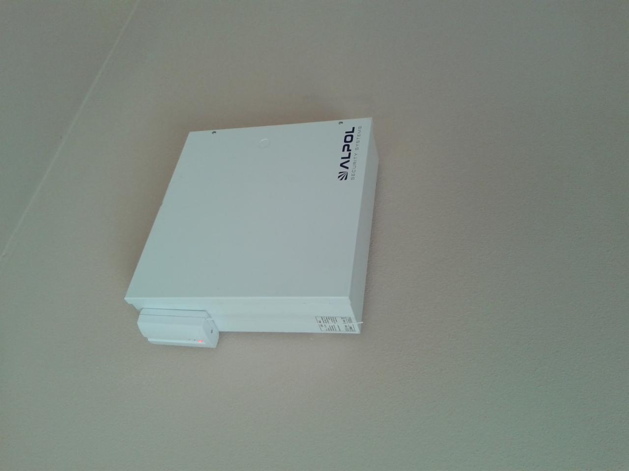 Biała centrala alarmowa firmy ALPOL SECURITY SYSTEMS z czujnikiem ruchu, zamontowana na jasnej ścianie wewnątrz budynku. Widok z dołu.