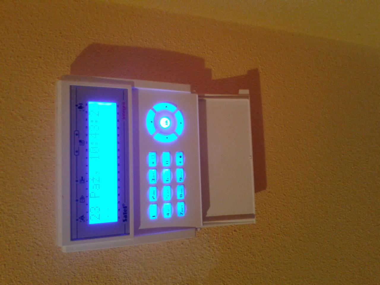 Biały panel sterowania alarmem Integra z niebieskim podświetleniem, zamontowany na żółtej ścianie. Widoczny ekran LCD i klawiatura numeryczna.