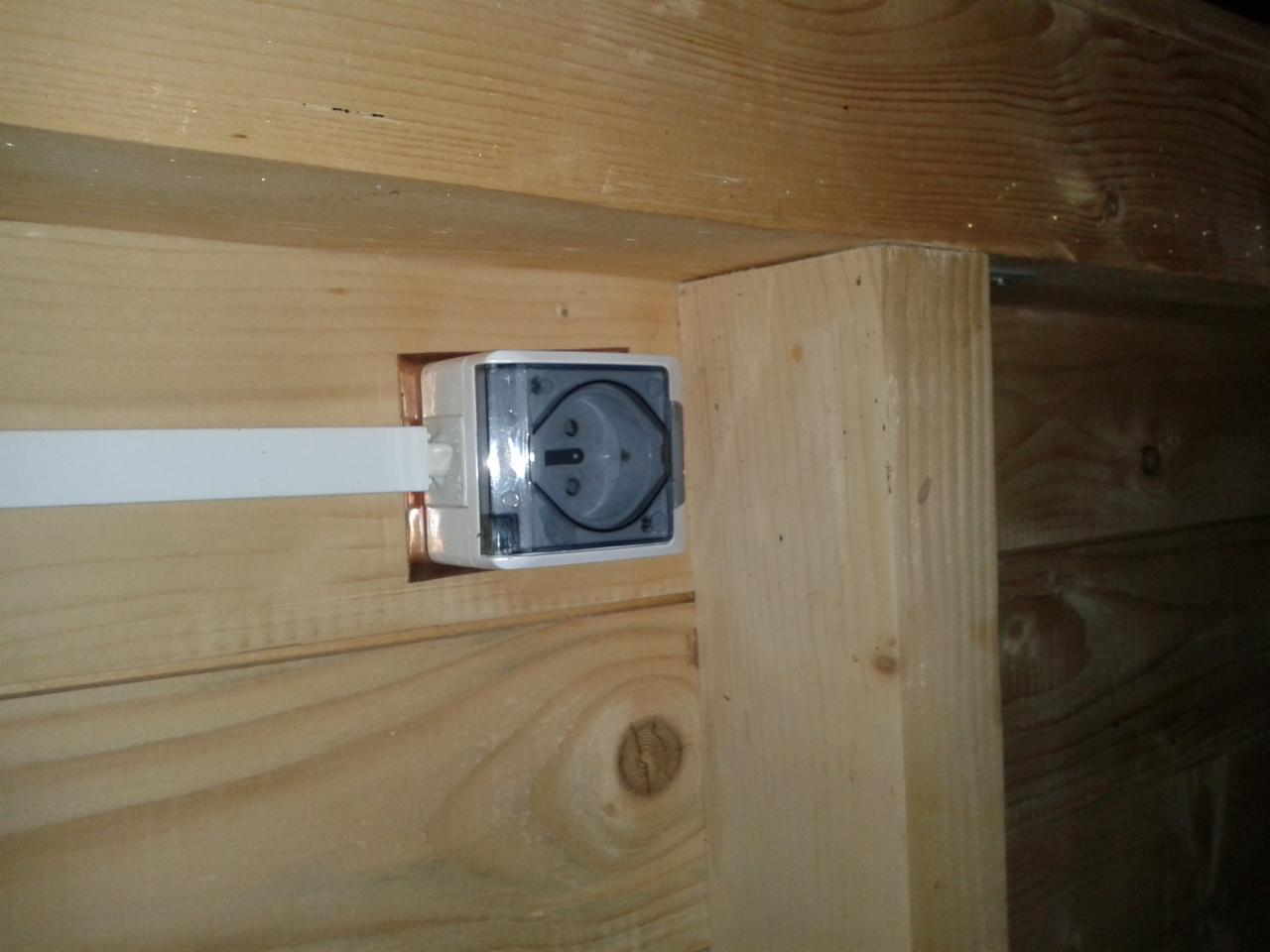 Zamontowane gniazdko elektryczne z przezroczystą osłoną w drewnianej ścianie. Widoczny kabel zasilający. Ujęcie z bliska na tle drewnianej konstrukcji.