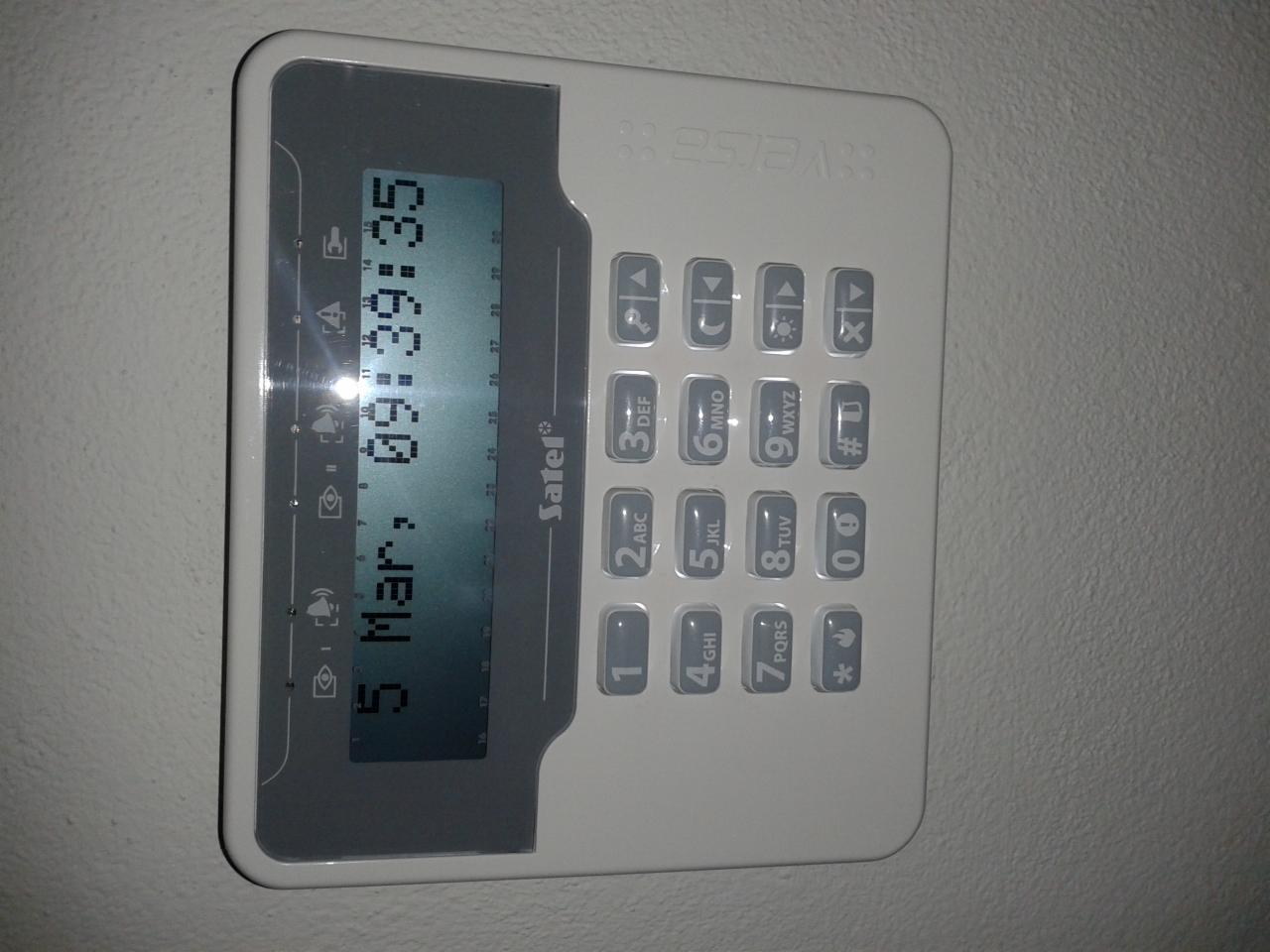 Panel sterowania alarmem Sater z wyświetlaczem LCD pokazującym datę i godzinę, umieszczony na jasnej ścianie. Widoczne przyciski numeryczne i funkcyjne.
