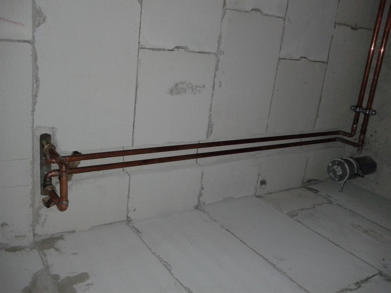 Miedziane rury centralnego ogrzewania zamontowane na ścianie z białych bloczków, widoczne złącza i fragment pompy obiegowej. Surowy stan, instalacja w trakcie.