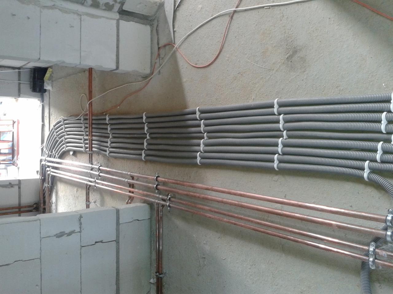 Szereg szarych, karbowanych rur i miedzianych rurek instalacyjnych, równolegle ułożonych na surowej ścianie w trakcie budowy, z widocznymi elementami mocującymi.