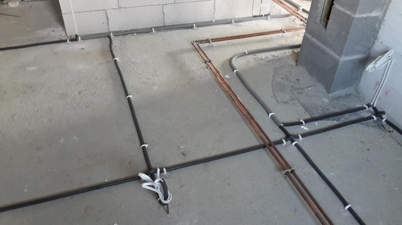 Instalacja podłogowa: rury miedziane i czarne peszle przymocowane do surowego betonu, przygotowane do zalania posadzką. Widoczny fragment ściany z betonu komórkowego.