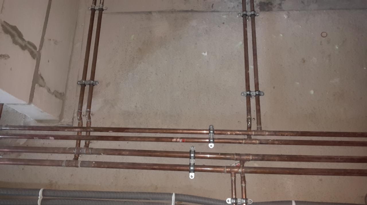 Miedziane rury instalacji centralnego ogrzewania, zamocowane do surowej ściany. Widoczne złączki i uchwyty mocujące. Szare rury karbowane w dolnej części kadru.