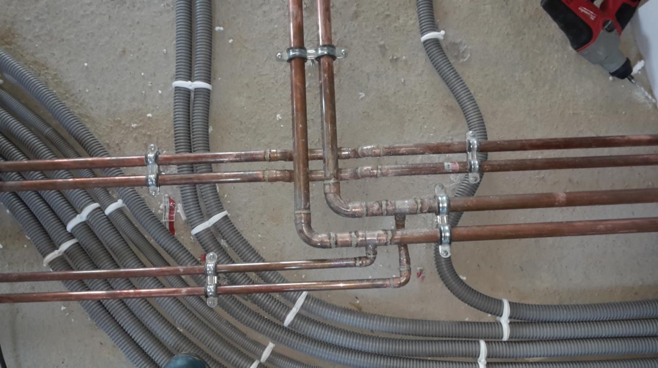 Miedziane rury instalacji CO i szare rury karbowane ułożone na surowym betonie, zamocowane uchwytami. W tle wiertarka.