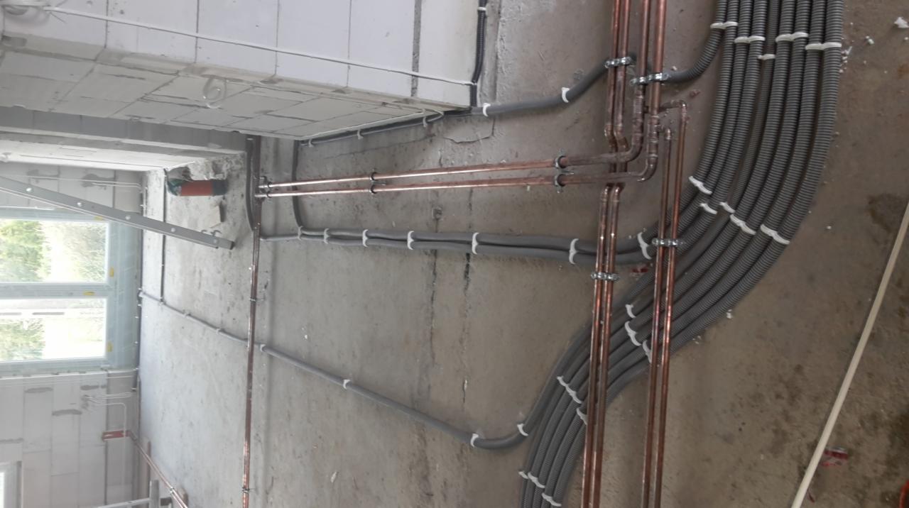 Miedziane i szare rury instalacyjne prowadzone wzdłuż surowej ściany, z widocznymi mocowaniami i fragmentem okna. Instalacje w nowym budynku.