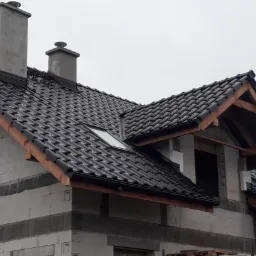 Nowy, czarny dach z dachówki ceramicznej na budynku w trakcie budowy, widoczne kominy i okno dachowe.