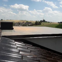 Widok z góry na dwa rodzaje pokrycia dachowego: dachówka ceramiczna w pierwszym planie i płaski dach z papy termozgrzewalnej w tle, z kominami i widokiem na wiejską zabudowę oraz pola uprawne...