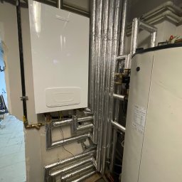 Atlantis Technika Grzewcza i Sanitarna Piotr Szajewski - Biały piec gazowy marki Brotje zamontowany na ścianie, z widocznymi rurami w srebrnej izolacji, obok stoi biały zasobnik ciepłej wody marki Beretta.