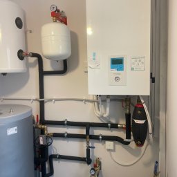 Atlantis Technika Grzewcza i Sanitarna Piotr Szajewski - Biały wiszący piec gazowy Panasonic Aquarea z podłączonymi rurami, zbiornikami i osprzętem, zamontowany na ścianie w pomieszczeniu technicznym.