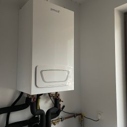Atlantis Technika Grzewcza i Sanitarna Piotr Szajewski - Nowy, biały, wiszący kondensacyjny kocioł gazowy Brotje Heizung z podłączonymi, zaizolowanymi rurami instalacji grzewczej, widoczne zawory i przyłącza, zamontowany na tle białej ściany...