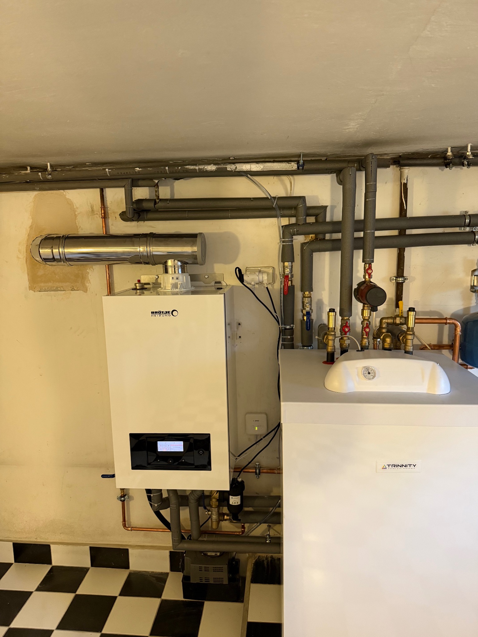 Biały piec gazowy z chromowanym kominem i rozbudowaną instalacją hydrauliczną, widoczne liczne rury miedziane i zawory, na ścianie widoczne ślady po malowaniu.