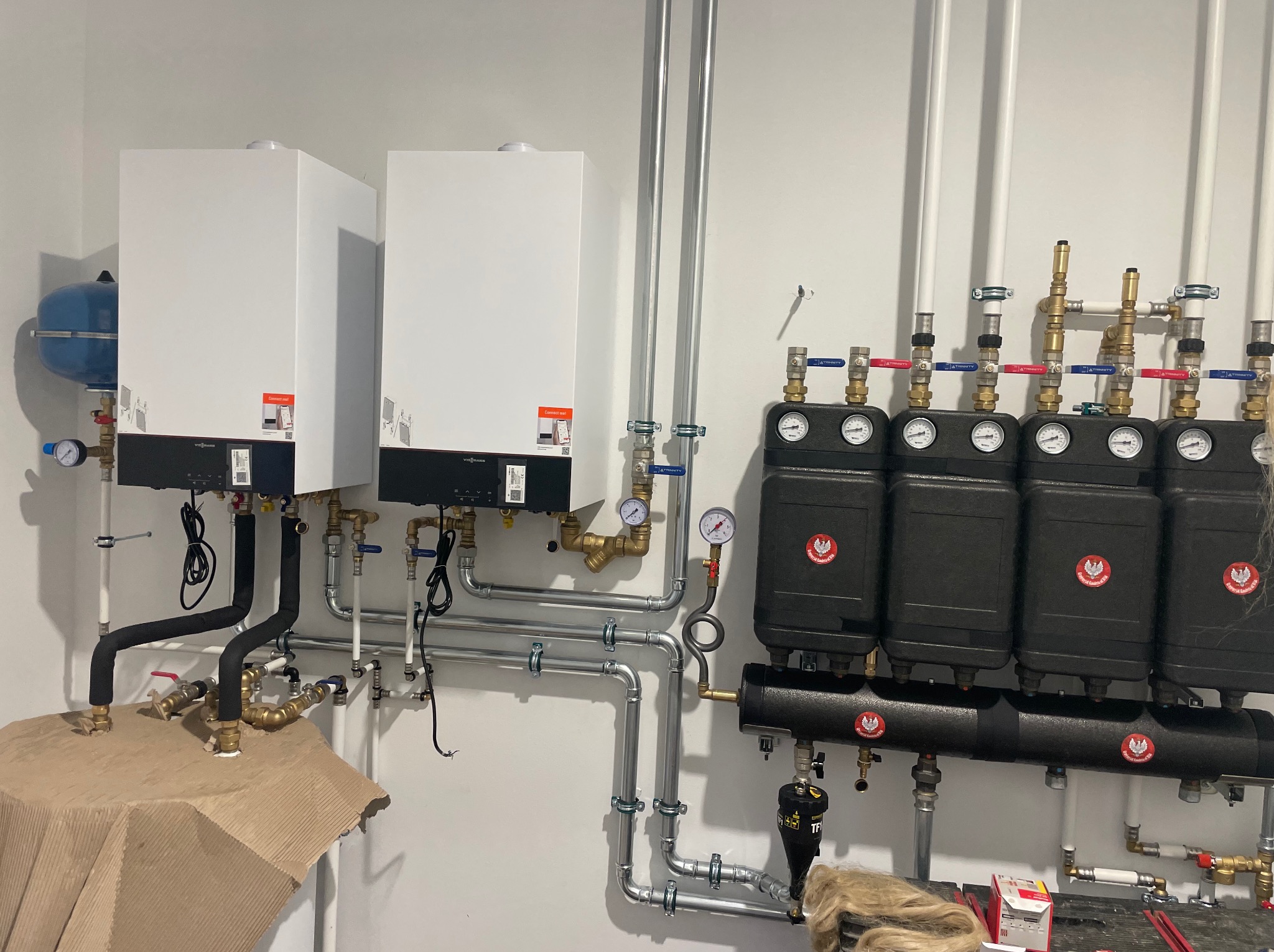 Dwa białe kotły gazowe Viessmann zamontowane na ścianie, z widocznymi rurami i zaworami, oraz cztery czarne rozdzielacze z manometrami, całość instalacji grzewczej w trakcie montażu, zabezpieczona...