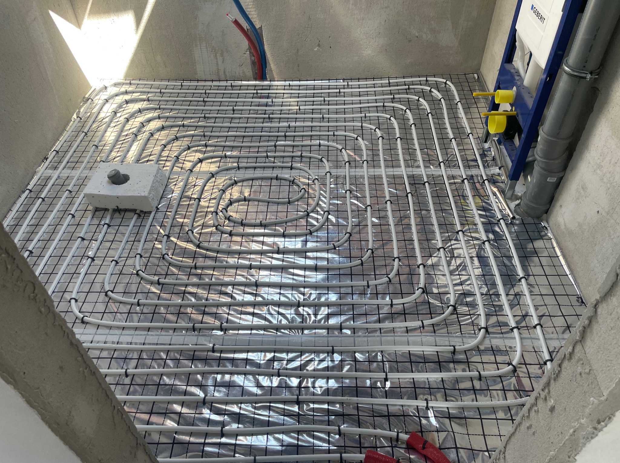 Instalacja ogrzewania podłogowego w surowym wnętrzu, rury grzewcze ułożone na folii refleksyjnej z czarną siatką, widoczny fragment ściany z instalacją sanitarną Geberit.