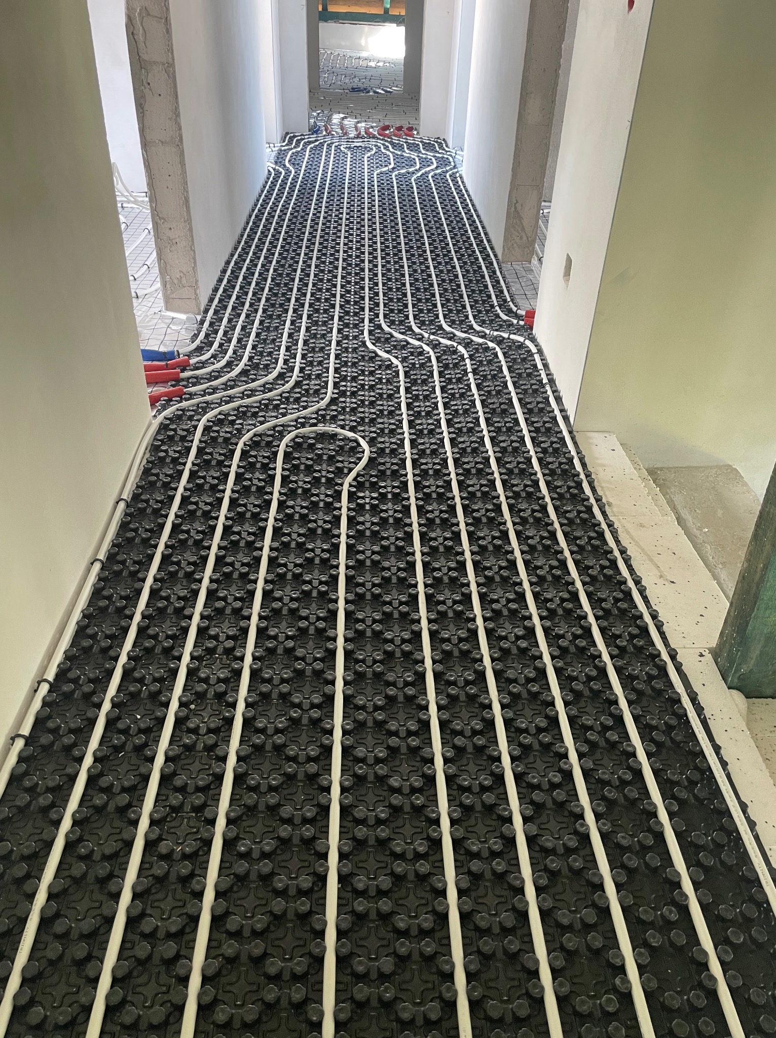 Białe rury grzewcze ułożone w pętlach na czarnej, wypustkowej macie w trakcie montażu ogrzewania podłogowego w surowym wnętrzu.