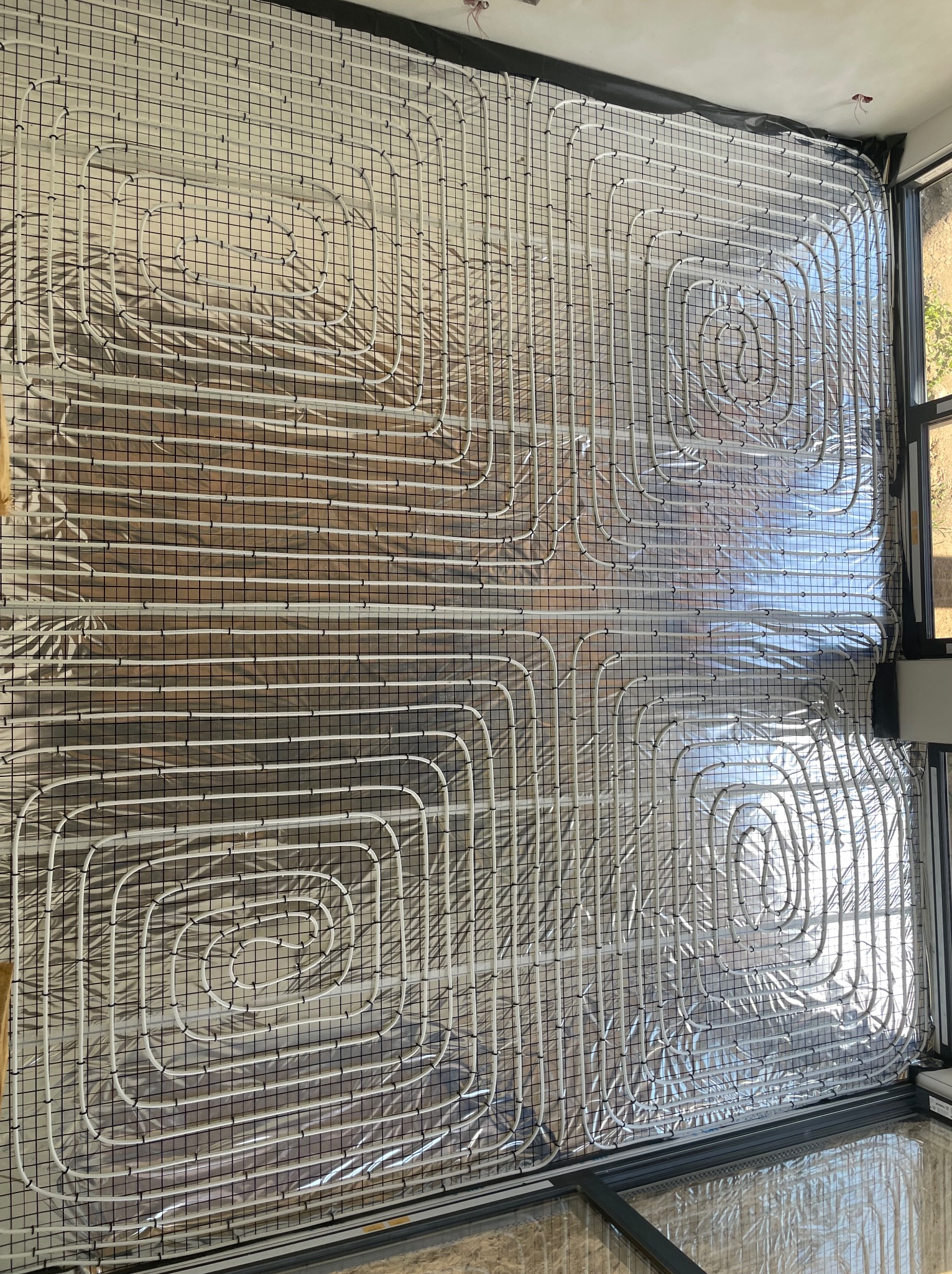 Rozłożona instalacja ogrzewania podłogowego na folii izolacyjnej, przytwierdzona do metalowej siatki, widok z góry, przygotowanie do wylewki.