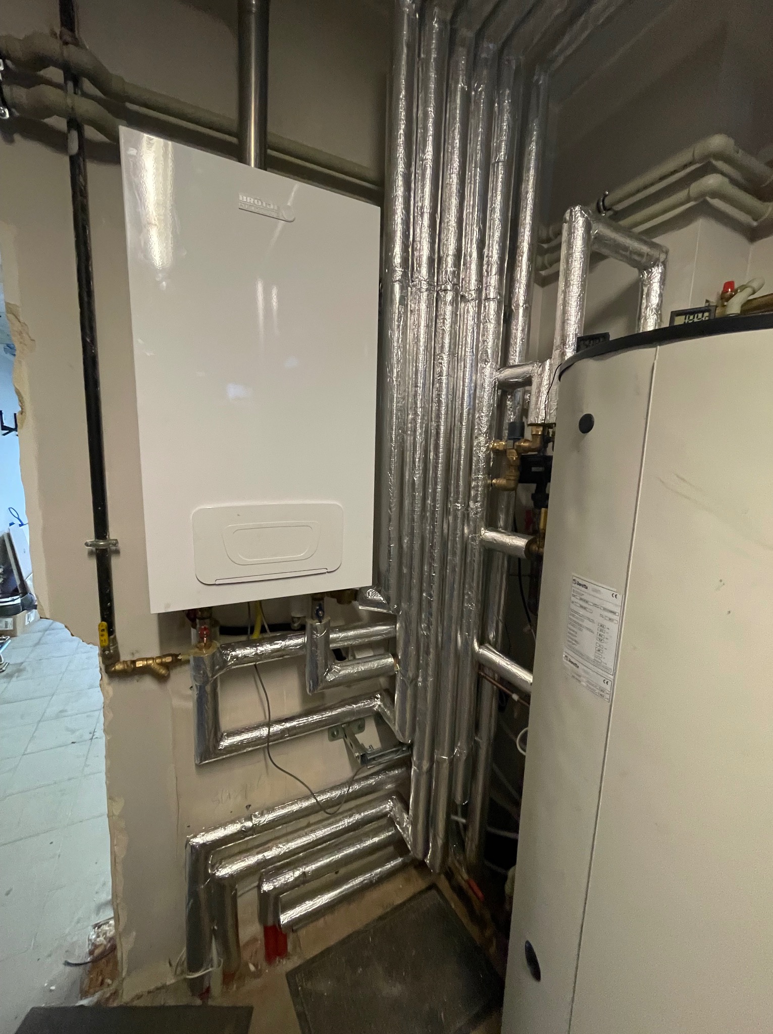 Biały piec gazowy marki Brotje zamontowany na ścianie, z widocznymi rurami w srebrnej izolacji, obok stoi biały zasobnik ciepłej wody marki Beretta.