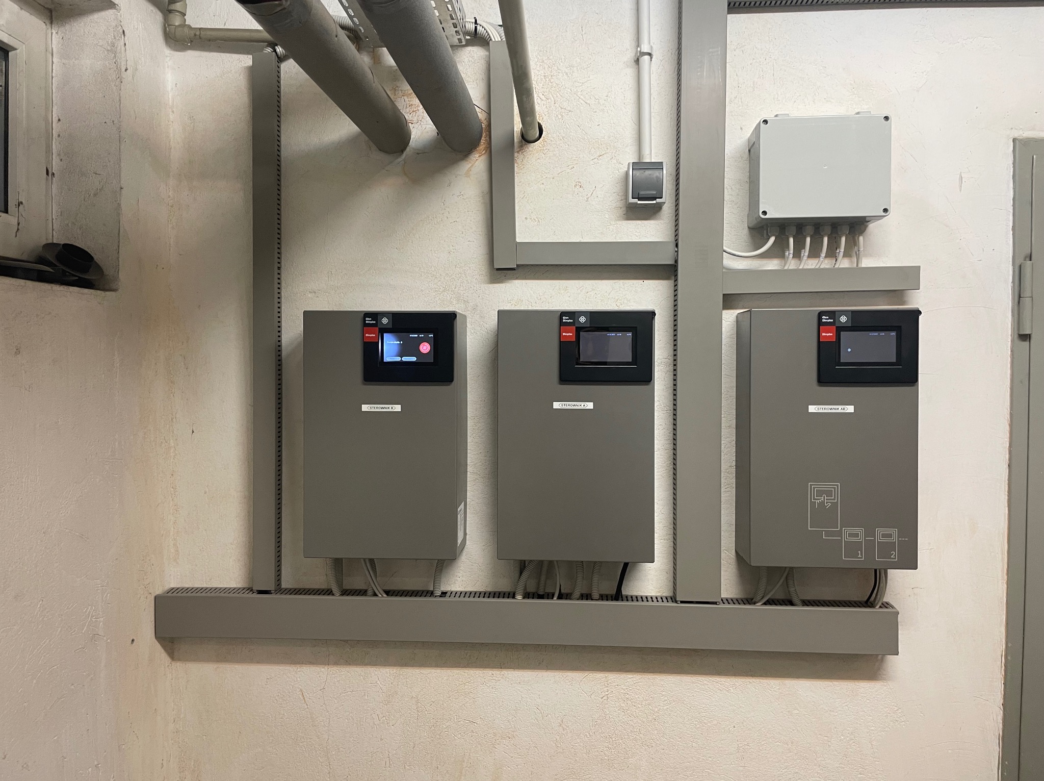 Trzy szare panele sterowania z ekranami dotykowymi zamontowane na ścianie w pomieszczeniu technicznym, z widocznymi rurami instalacyjnymi i skrzynką elektryczną.