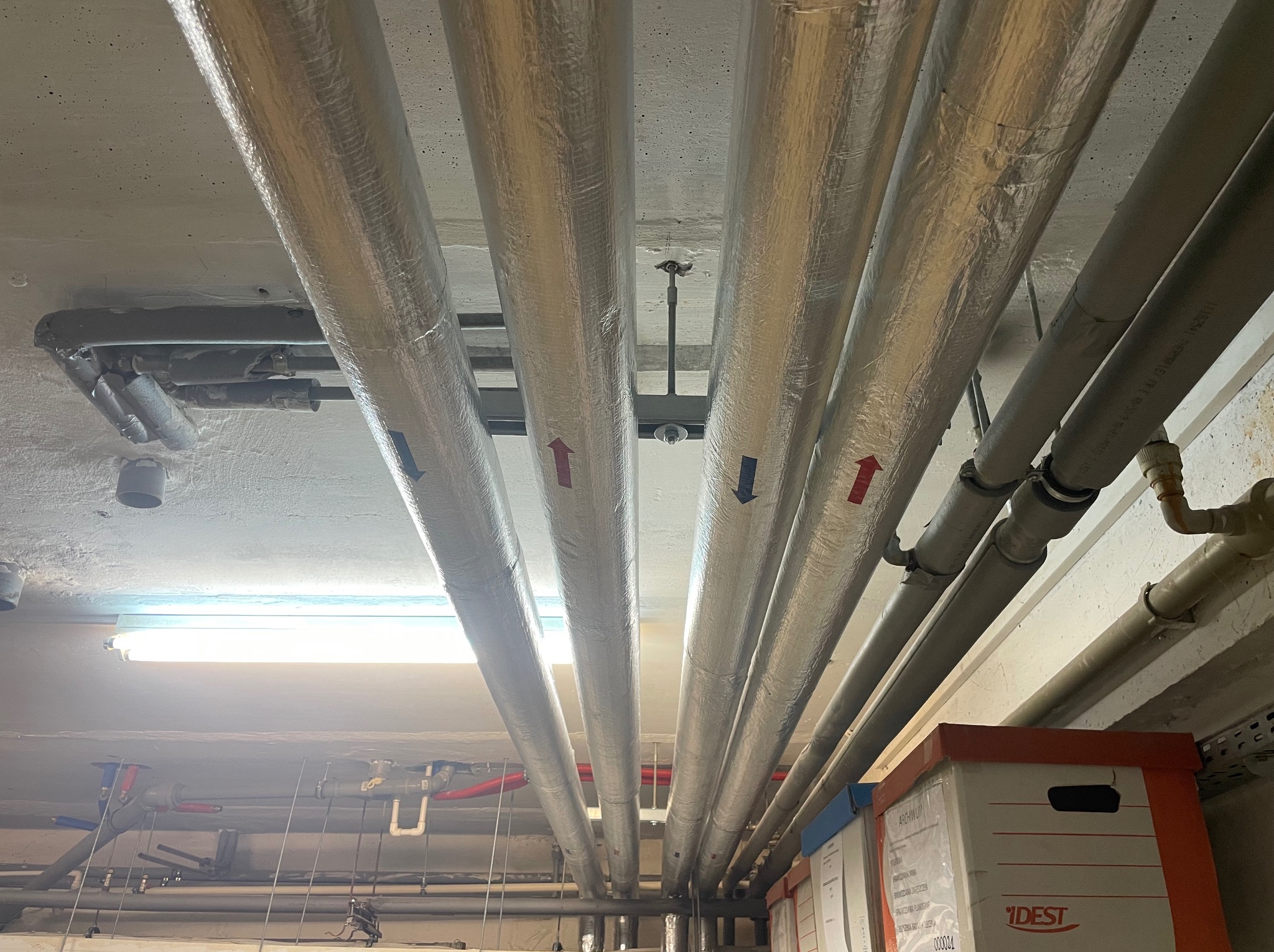 Rury instalacji grzewczej, zaizolowane folią aluminiową, biegnące pod sufitem, z czerwonymi i niebieskimi strzałkami oznaczającymi kierunek przepływu, widok z dołu, w tle widoczne inne elementy...