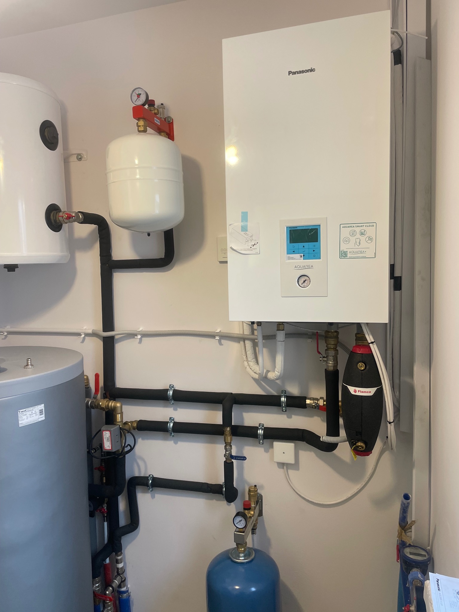 Biały wiszący piec gazowy Panasonic Aquarea z podłączonymi rurami, zbiornikami i osprzętem, zamontowany na ścianie w pomieszczeniu technicznym.