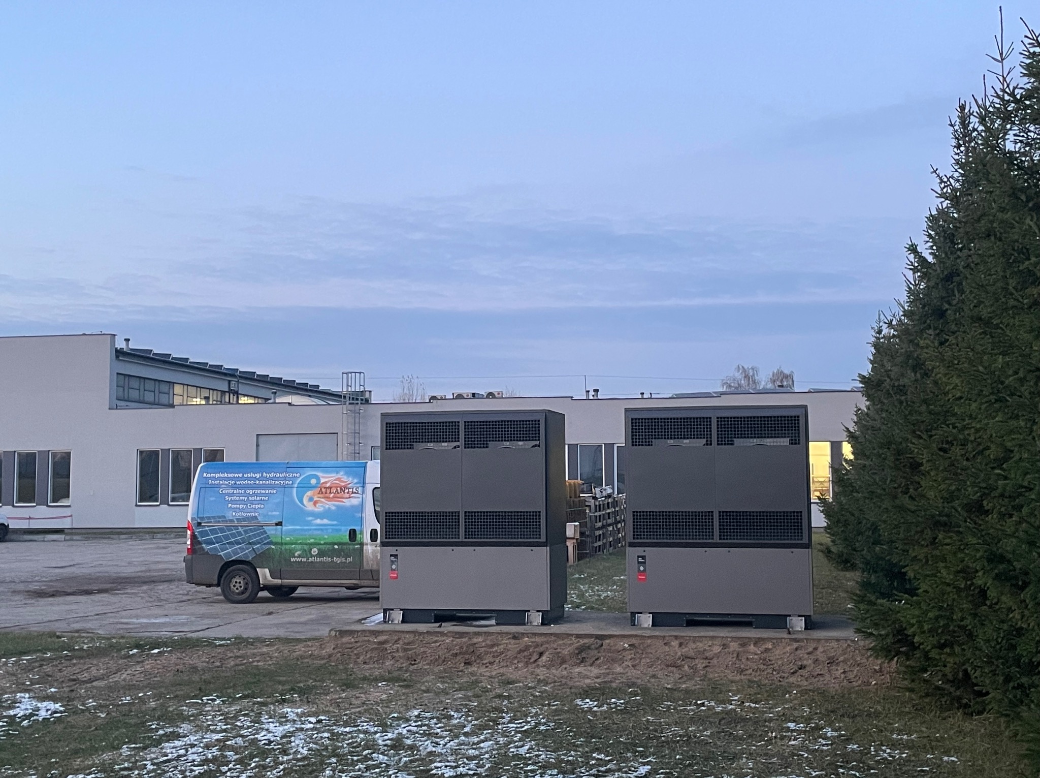 Dwa duże, szare urządzenia grzewcze ustawione na zewnątrz budynku, obok firmowego samochodu dostawczego z reklamą usług hydraulicznych i grzewczych. Na ziemi resztki śniegu, w tle budynek i drzewa...