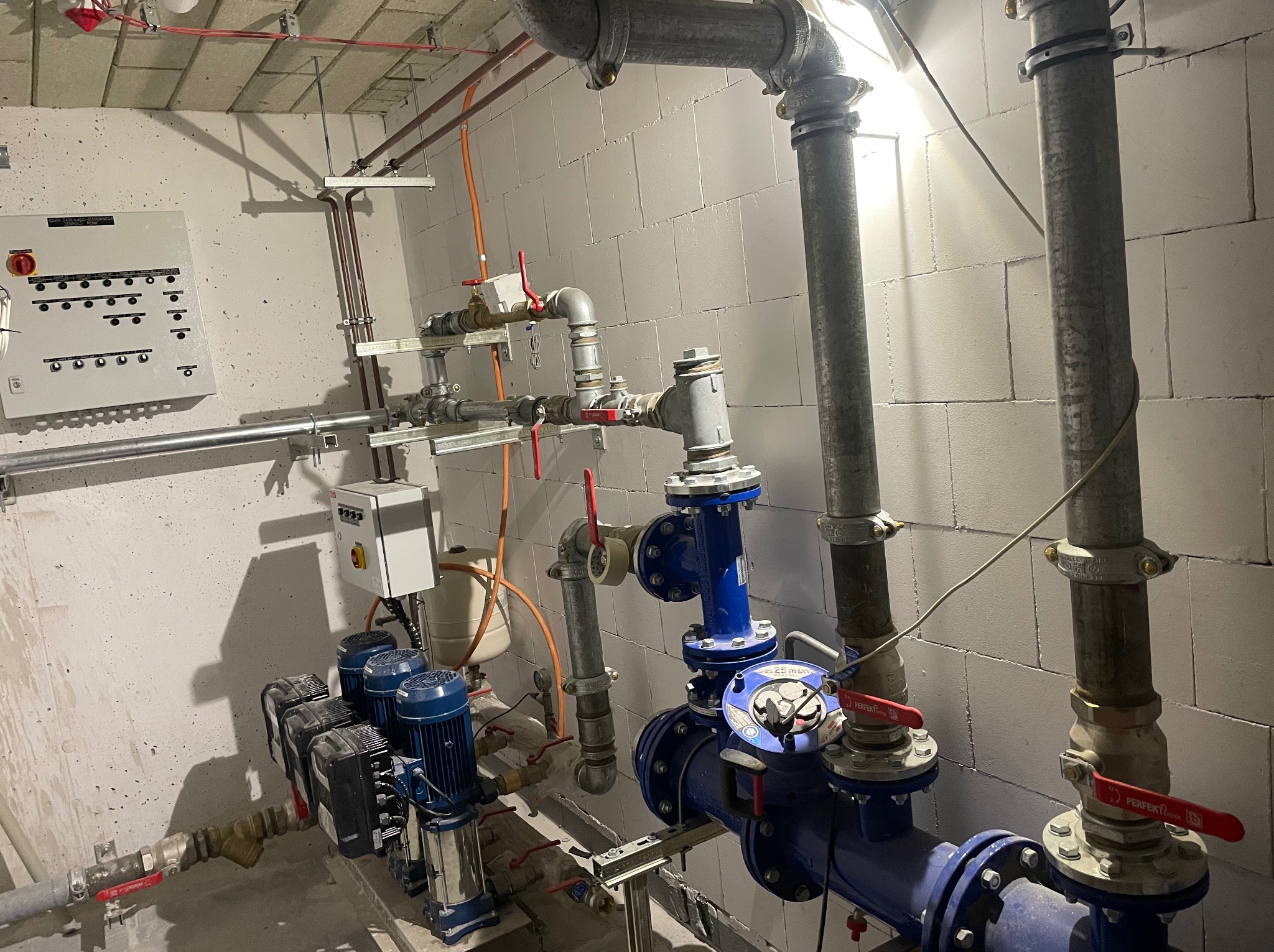 Węzeł instalacyjny z pompami, rurami, zaworami i szafą sterowniczą na tle ściany z bloczków betonowych. Widoczne liczne połączenia gwintowane i kołnierzowe.
