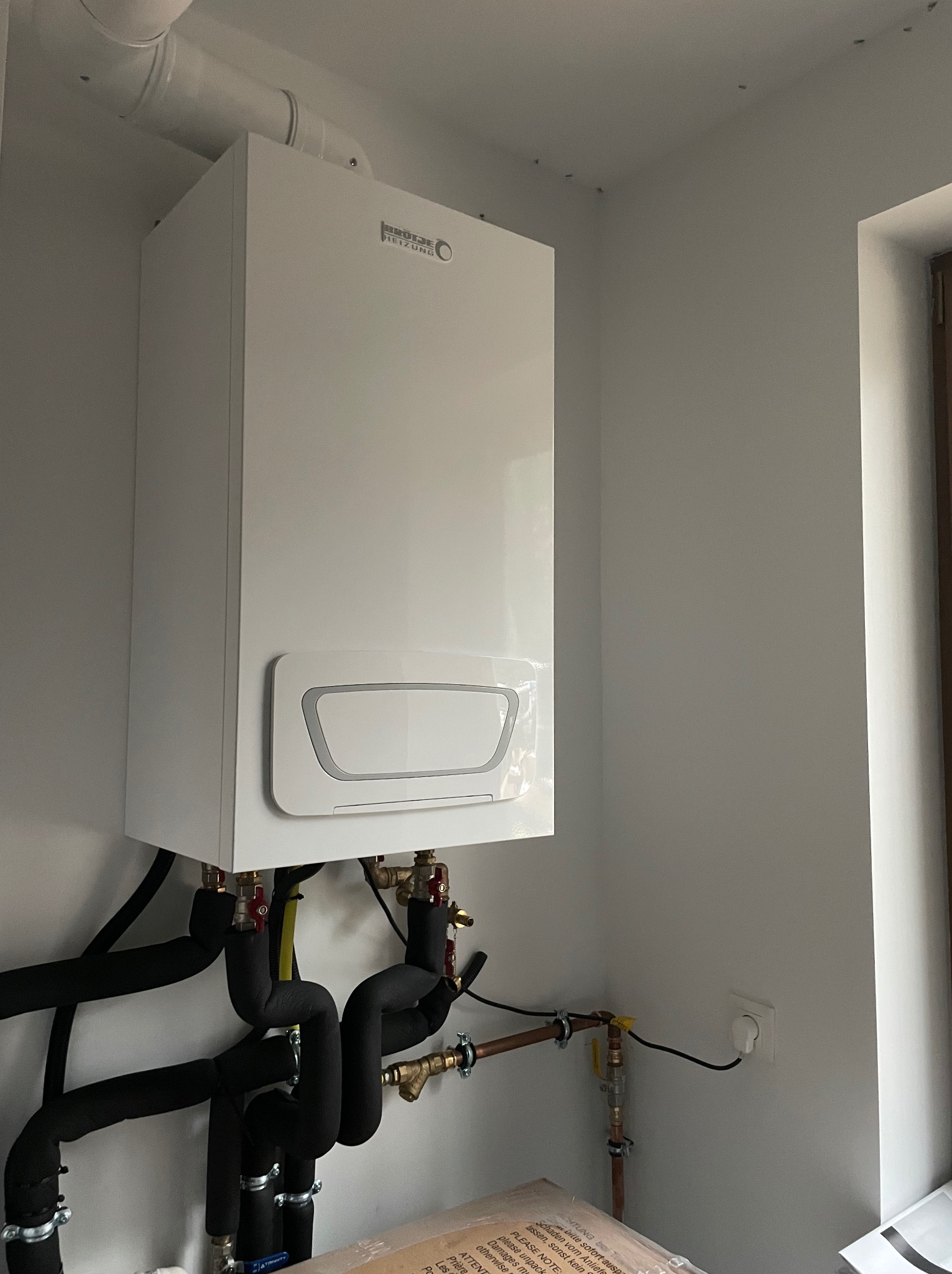 Nowy, biały, wiszący kondensacyjny kocioł gazowy Brotje Heizung z podłączonymi, zaizolowanymi rurami instalacji grzewczej, widoczne zawory i przyłącza, zamontowany na tle białej ściany...