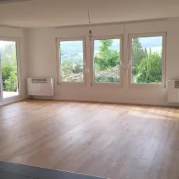 Jasne, przestronne wnętrze z panelami podłogowymi w odcieniach drewna i ciemnymi, grafitowymi płytkami w przejściu, trzy okna z widokiem na zieleń, białe ściany i grzejniki.