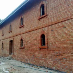 Ceglana ściana dwukondygnacyjnego budynku z łukowymi oknami, widoczne ślady prac budowlanych i gruz na ziemi.