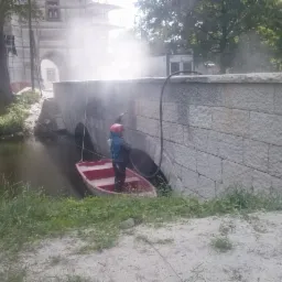 Pracownik w czerwonym kasku i niebieskiej kamizelce, stojący w czerwonej łódce na rzece, czyści kamienny mur mostu za pomocą strumienia pod ciśnieniem. W tle widać budynek w rusztowaniach i drzewa.